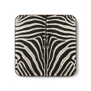 May include: A square coaster featuring a black and white zebra stripe pattern. The surface displays a symmetrical arrangement of bold, dark stripes against a cream-colored background, finished with a thin, dark brown border along the edges.