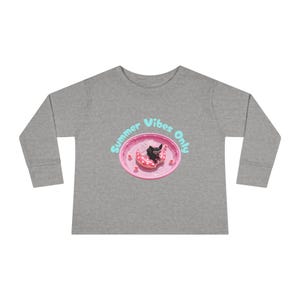 Peut inclure: Un t-shirt gris chiné à manches longues avec un motif représentant un petit chien noir portant des lunettes de soleil roses, flottant sur une bouée rose à pois dans une piscine rose. Le texte Summer Vibes Only est imprimé en bleu clair.