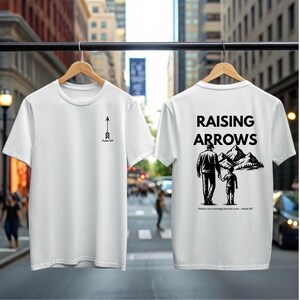 Könnte beinhalten: Zwei weiße T-Shirts hängen an einem Gestell. Eines hat ein schwarzes Pfeildesign mit dem Text "Psalm 127". Das andere Shirt zeigt die Worte "RAISING ARROWS" über einer Grafik eines Elternteils und Kindes vor Bergen.