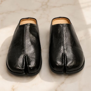 May include: A pair of black slip-on shoes with a distinct split-toe design. The shoes feature a smooth, slightly textured leather-like finish, a central seam running down the front, and a tan-colored interior lining visible at the open heel area.