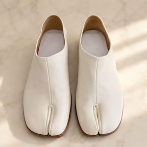 May include: A pair of off-white split-toe shoes featuring a smooth, matte finish. The footwear has a distinct vertical seam running down the center of the vamp, a low-cut topline, and a light-colored interior lining with a flat, brown sole.