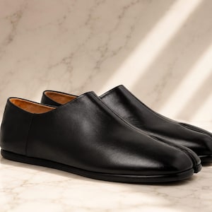 Pode incluir: Um par de sapatos sem atacadores em pele preta com um design distinto de biqueira dividida. Os sapatos têm um acabamento liso e mate, uma silhueta de perfil baixo e um forro interior bege, exibindo uma construção limpa sem atacadores.