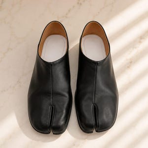 May include: A pair of black leather slip-on shoes featuring a distinct split-toe design. The shoes have a smooth black exterior, a tan interior lining, and a white insole. The construction includes a central seam running down the front.