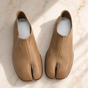 May include: A pair of tan leather tabi shoes featuring a distinct split-toe design. The shoes have a smooth, matte finish, a high-cut ankle opening, and a light-colored interior lining. The construction includes a central seam running down the front.