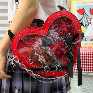 Può includere: Una borsa a tracolla rossa a forma di cuore con una finestra frontale trasparente, ali di pipistrello nere e un dettaglio con nastro nero. La borsa è decorata con borchie argentate, una catena argentata con gemme rosse e rose rosse.