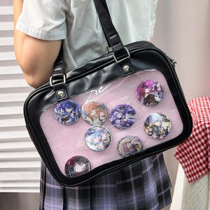 Könnte beinhalten: Eine schwarze, rechteckige Umhängetasche mit einem großen, durchsichtigen Sichtfenster auf der Vorderseite, in dem sieben runde Anstecknadeln mit Anime-Motiven auf einem hellrosa Stoffeinsatz befestigt sind. Die Tasche hat schwarze Kunstlederbesätze.