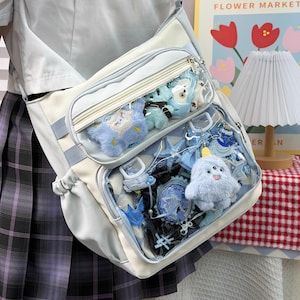 Könnte beinhalten: Eine hellblaue und weiße Ita-Tasche mit zwei durchsichtigen Vordertaschen, die mit verschiedenen blauen Plüschanhängern, kleinen Figuren und dekorativen Accessoires gefüllt sind. Die Tasche hat einen Reißverschluss und verstellbare Trageriemen.