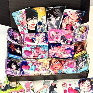 May include: A black box filled with various rectangular acrylic charms and stickers featuring colorful anime-style illustrations of characters with blushing expressions, hearts, and the text Alienstage, arranged in a dense, overlapping collection.