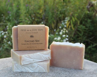 Op de afbeelding: Vier staven natuurlijke, handgemaakte zeep gestapeld op een grijs oppervlak. De bovenste staaf heeft een bruin papieren etiket met de tekst "Fresh as a Daisy Soaps" en "Rosemary Mint Shampoo Bar with Sweet & Shea Butter Net Wt. 2.5 oz / 71 g".