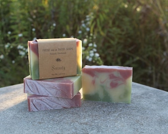 May include: A stack of handmade soaps with a natural aesthetic. The soaps feature layers of green, pink, and white, with one wrapped in brown paper labeled "Serenity." The background is blurred greenery.
