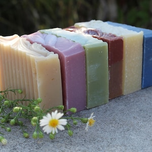 May include: Six bars of handmade soap in various colors, including lavender, green, brown, and blue. The bars are stacked on top of each other and are arranged in a rainbow pattern. The soap is sitting on a gray surface.