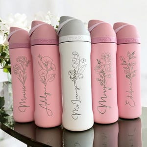 Könnte beinhalten: Fünf Owala-Wasserflaschen in einer Reihe. Vier Flaschen sind mattrosa mit hellrosa Deckeln, eine mittlere Flasche ist weiß mit grauem Deckel. Jede Flasche zeigt eine florale Linienzeichnung und einen individuellen Namen in Schreibschrift.