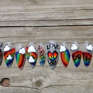 Pode incluir: Um conjunto de dez unhas artificiais com uma base prateada brilhante. Cada unha apresenta um arco-íris, nuvens brancas e as palavras "LOVE IS LOVE" e "PRIDE" em letras pretas. As unhas estão dispostas sobre uma superfície de madeira.