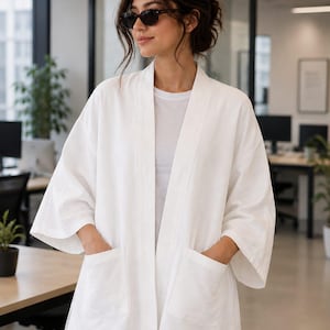 Puede incluir: Una chaqueta blanca de estilo kimono abierta por delante, con mangas tres cuartos, cuello sin solapas y dos grandes bolsillos de parche en la parte delantera. La prenda se lleva sobre una camiseta blanca lisa y unos vaqueros azul claro.
