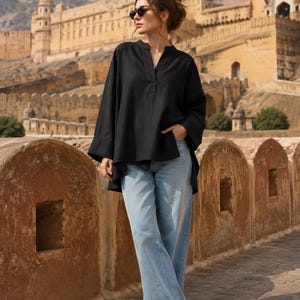 Könnte beinhalten: Eine schwarze Langarmtunika mit geschlitztem Ausschnitt und asymmetrischem Saum, kombiniert mit einer hellblauen, weit geschnittenen Jeans und weißen Turnschuhen. Die Tunika hat eine lockere Passform mit weiten Ärmeln und ein schlichtes Design.