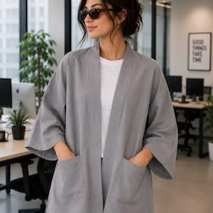 Puede incluir: Una chaqueta gris abierta por delante con mangas tres cuartos y dos grandes bolsillos de parche en la parte frontal. La prenda se lleva sobre una camiseta blanca lisa y vaqueros azul claro, con un diseño sencillo sin cuello y aberturas.