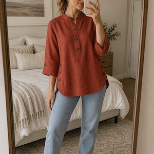 May include: A rust-colored, three-quarter sleeve tunic shirt featuring a mandarin collar, a partial front button placket, and side slits with decorative buttons, paired with light blue denim jeans and white sneakers.
