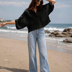 May include: A black short-sleeved button-down shirt with a textured finish paired with light blue denim jeans and white trainers. The shirt features a loose, boxy fit with wide sleeves, while the jeans have a straight-leg cut and a classic five-pocket design.
