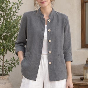May include: A charcoal gray jacket featuring a mandarin collar, three-quarter length sleeves with folded cuffs, and five light-colored wooden buttons down the front. The jacket includes two front patch pockets and is worn over a white top.