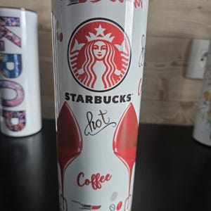 Peut inclure: Un gobelet cylindrique blanc orné du logo Starbucks, du mot "hot" en écriture cursive, du mot "Coffee" en rouge, ainsi que de motifs de chaussures à talons rouges, de rouges à lèvres et de grains de café. Il possède un couvercle transparent.