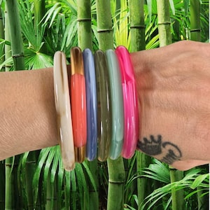 Peut inclure: Un ensemble de six bracelets joncs cylindriques et lisses de différentes couleurs, notamment crème, corail, bleu clair, vert olive, vert menthe et rose vif, portés ensemble sur un poignet devant un arrière-plan de tiges de bambou vert.