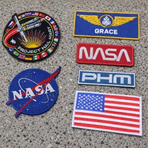 May include: A collection of six embroidered patches including a circular Project Hail Mary patch with international flags, a blue Grace patch with gold wings, a red NASA patch, a blue PHM patch, a circular NASA logo patch, and a rectangular American flag.