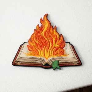 May include: An embroidered patch featuring an open book with pages rendered in cream and brown thread. A large, vibrant flame in shades of yellow, orange, and red rises from the center of the pages. A small green bookmark hangs from the bottom edge.