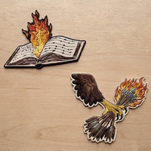 May include: Two embroidered patches. One patch depicts an open book with flames rising from the pages. The second patch shows a bird of prey with brown wings and yellow talons clutching a bundle of arrows engulfed in bright orange and yellow flames.