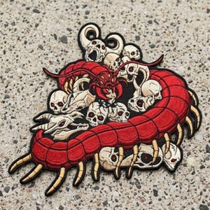 May include: An embroidered patch featuring a large, segmented red centipede winding through a cluster of off-white skulls with red eye sockets. The centipede has numerous tan legs and a dark red head, while the skulls are arranged in a dense pile.