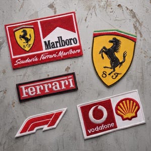 May include: A collection of five embroidered patches featuring racing logos. Included are a rectangular Scuderia Ferrari Marlboro patch, a shield-shaped Ferrari prancing horse emblem, a rectangular Ferrari logo, a red F1 logo, and a Vodafone Shell patch.
