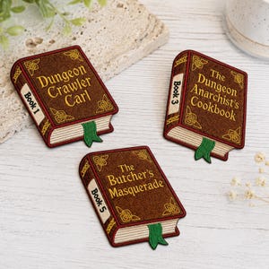 May include: Three rectangular embroidered patches shaped like books with brown covers, gold lettering, and green bookmarks. The titles read Dungeon Crawler Carl Book 1, The Dungeon Anarchist's Cookbook Book 3, and The Butcher's Masquerade Book 5.