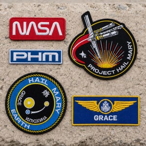 May include: A collection of five embroidered patches including a red NASA rectangle, a blue PHM rectangle, a circular Project Hail Mary patch with a spacecraft, a circular Hail Mary Earth Grace patch with Cyrillic text, and a blue Grace wing patch.