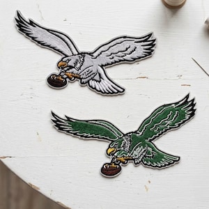May include: Two embroidered fabric patches featuring eagles in flight, each clutching a brown football in its talons. One eagle is rendered in shades of silver and white, while the other is depicted in dark green and white with yellow beaks.