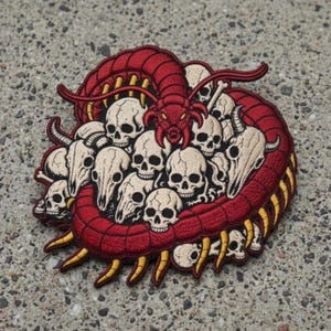 May include: An embroidered patch featuring a large red centipede coiled around a cluster of beige human and animal skulls. The centipede has yellow legs and sharp mandibles, while the skulls are detailed with black eye sockets and nasal cavities.
