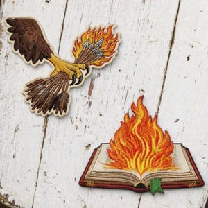 May include: Two embroidered patches. One patch depicts a brown eagle with yellow talons clutching a bundle of arrows surrounded by orange and yellow flames. The second patch shows an open book with a large, vibrant orange and yellow flame rising from the pages, featuring a small green bookmark at the bottom.
