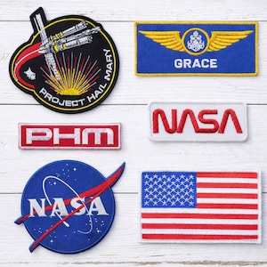May include: Six embroidered patches including a circular Project Hail Mary patch, a blue Grace wing patch, a rectangular PHM patch, a white NASA patch, a circular blue NASA meatball patch, and a rectangular United States flag patch.