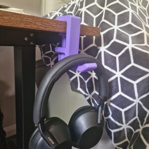 May include: A matte purple C-clamp headphone hanger attached to the edge of a wooden desk, holding a pair of black Sony over-ear headphones. The hanger features a threaded screw mechanism for tightening against the desk surface.
