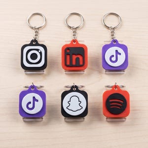 Könnte beinhalten: Sechs quadratische NFC-Social-Media-Schlüsselanhänger mit Metallringen, die Logos von Instagram, LinkedIn, TikTok, Snapchat und Spotify zeigen. Der Text SMART NFC SOCIAL TAGS steht in schwarzer Schrift über den bunten Silikonanhängern.