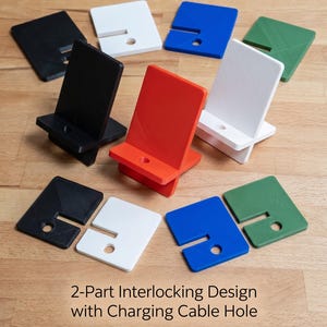 May include: A collection of 2-part interlocking phone stands in black, white, red, blue, and green. Each stand features a flat base and an upright support with a circular hole for a charging cable. The text 2-Part Interlocking Design with Charging Cable Hole is visible.