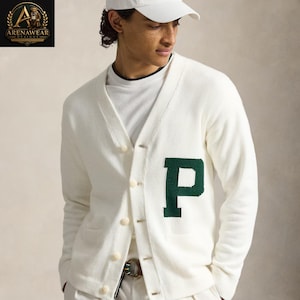 May include: Cream-colored cardigan sweater with a large green "P" on the front. The sweater has a V-neck, button closure, and two front pockets. The model is wearing a white baseball cap and a white t-shirt.