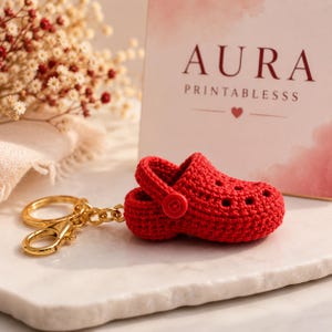 May include: A red crocheted clog-shaped keychain with a gold-colored chain and clasp. The keychain is next to a card that says "AURA PRINTABLESSS". The background includes flowers and a marble surface.