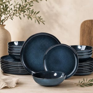 May include: A set of dark blue ceramic dinnerware, including plates and bowls, arranged on a wooden surface. The plates vary in size, with a textured finish. A vase with greenery is in the background.