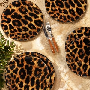 May include: A set of round dinner plates featuring a brown and black leopard print pattern, accompanied by a silver-toned metal fork and knife with brown handles. The plates are arranged in a group, showcasing the consistent animal print design.
