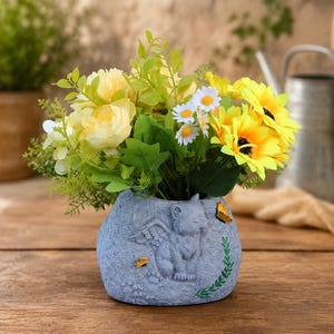 Puede incluir: Una maceta gris con textura de piedra que presenta un relieve de un gato alado con pequeños detalles de mariposas amarillas. La maceta contiene un arreglo de girasoles artificiales amarillos, margaritas blancas y follaje verde.
