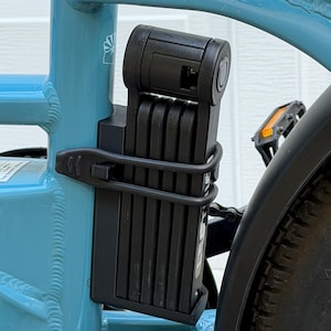May include: A black folding bicycle lock secured in a mounting bracket attached to the light blue frame of a bicycle. The lock consists of multiple rectangular steel segments connected by rivets, featuring a cylindrical locking mechanism at the top.