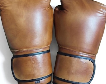 May include: A pair of tan leather boxing gloves with black trim. The left glove is labeled 16 oz and the right glove is labeled 12 oz. Both gloves feature a smooth, polished finish with visible stitching along the wrist and thumb areas.