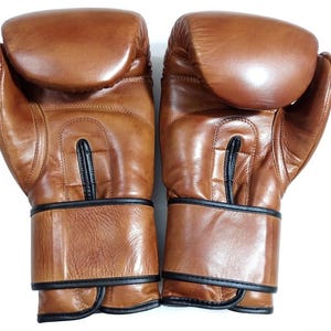 May include: A pair of tan leather boxing gloves featuring a smooth, rounded knuckle area, a central vertical stitched panel, and black piping trim along the wrist cuffs and seams. The gloves have a polished finish and a structured, padded design.