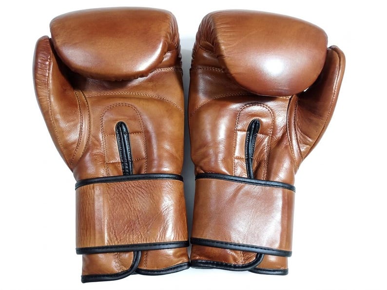 May include: A pair of tan leather boxing gloves featuring a smooth, rounded knuckle area, a central vertical stitched panel, and black piping trim along the wrist cuffs and seams. The gloves have a polished finish and a structured, padded design.