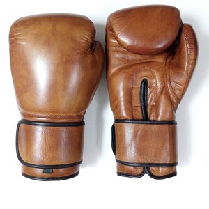 May include: A pair of tan leather boxing gloves with black trim and stitching. The left glove shows the text 12 OZ on a small label at the wrist. The gloves feature a smooth finish, a padded knuckle area, and a secure wrist strap closure.