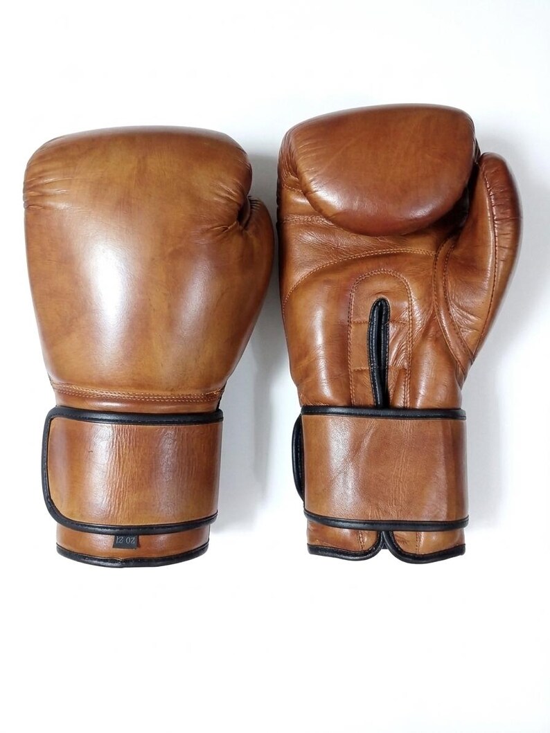 May include: A pair of tan leather boxing gloves with black trim and stitching. The left glove shows the text 12 OZ on a small label at the wrist. The gloves feature a smooth finish, a padded knuckle area, and a secure wrist strap closure.
